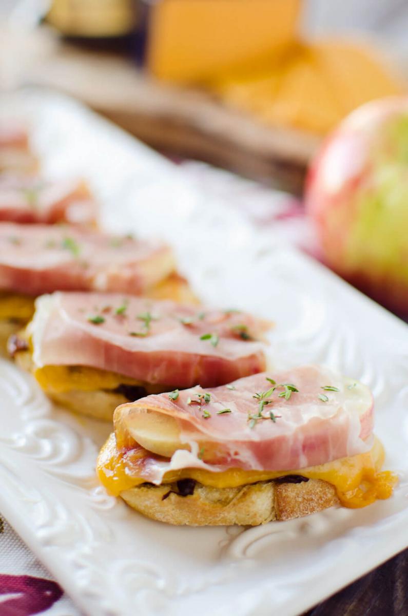 Caramelized onion and cheddar crostini with prosciutto wrapped apples on a white serving dish.