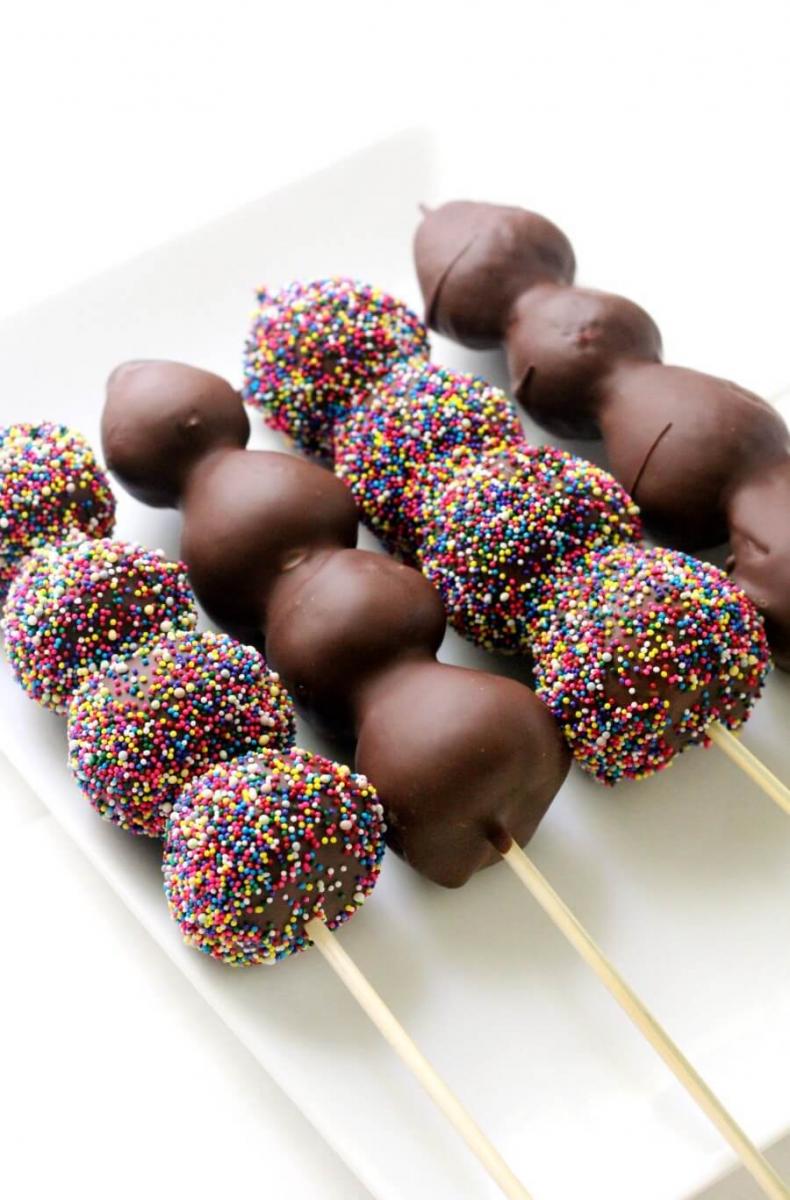 Four chocolate covered strawberry kebabs, two plain, two covered in rainbow sprinkles, on a white plate.  Each chocolate covered strawberry skewer has four berries.
