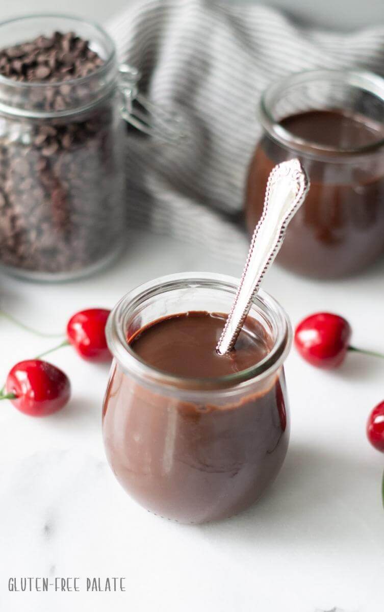 Glass jar of vegan hot fudge with a silver spoon sticking out, surrounded by fresh cherries.