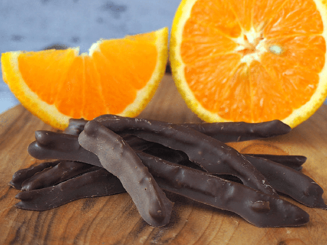Chocolate covered candied orange peels on a wooden cutting board in front of a sliced orange.