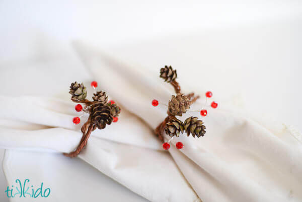 Two pinecone napkin rings with real pine cones and red crystals on white napkins.