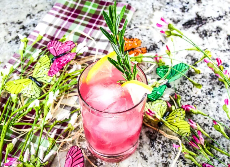 Bright pink citrus island guava cocktail garnished with lemon slices and fresh rosemary, surrounded by fresh wildflowers and yellow and pink silk butterflies.