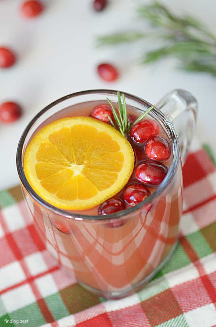 Glass mug filled with slow cooker cranberry cider, sitting on a red, white, and green plaid cloth.  An orange slice, fresh cranberries, and sprig of rosemary float in the mulled cider.