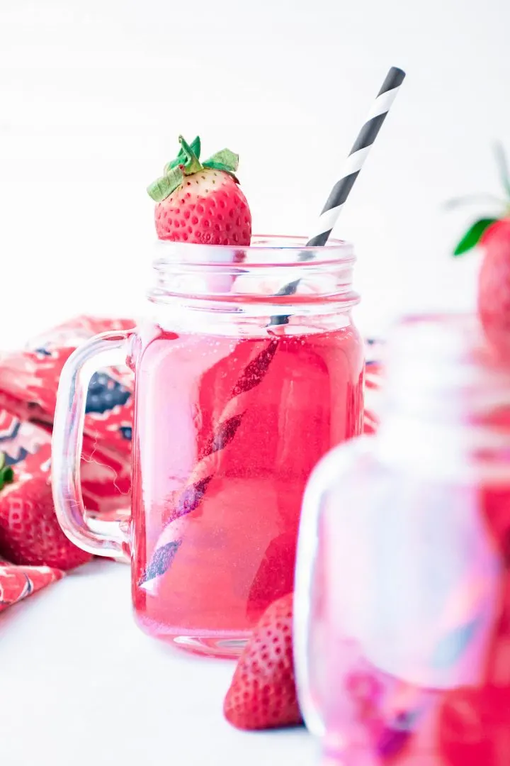 Mason jar glass filled with a bright pink Cupid's Cocktail recipe, garnished with a fresh strawberry and a black and white striped paper straw.