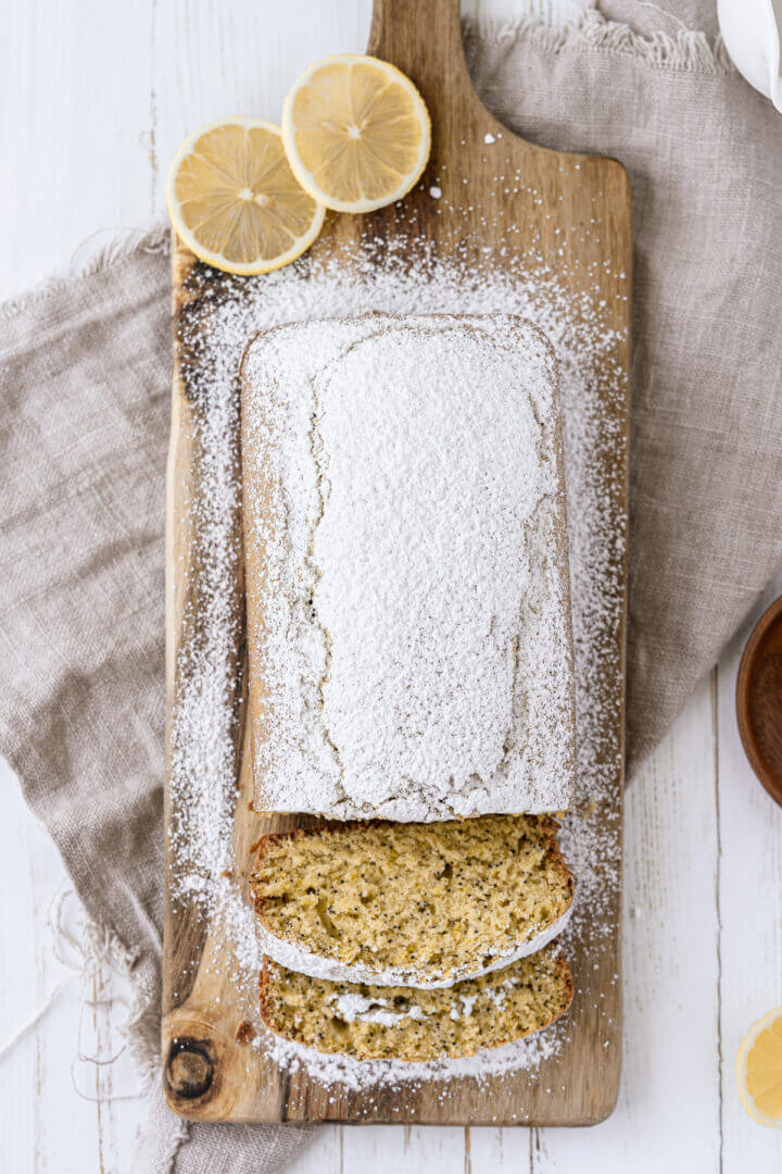 Vegan lemon poppy seed loaf dusted with powdered sugar and sliced on a wooden cutting board.