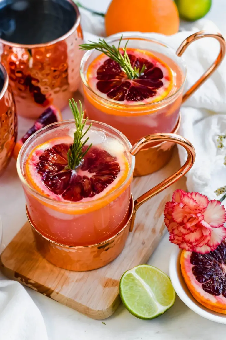 Blood orange moscaw mule cocktails in a copper-bottomed glass, garnished with a slice of blood orange and a sprig of fresh rosemary.