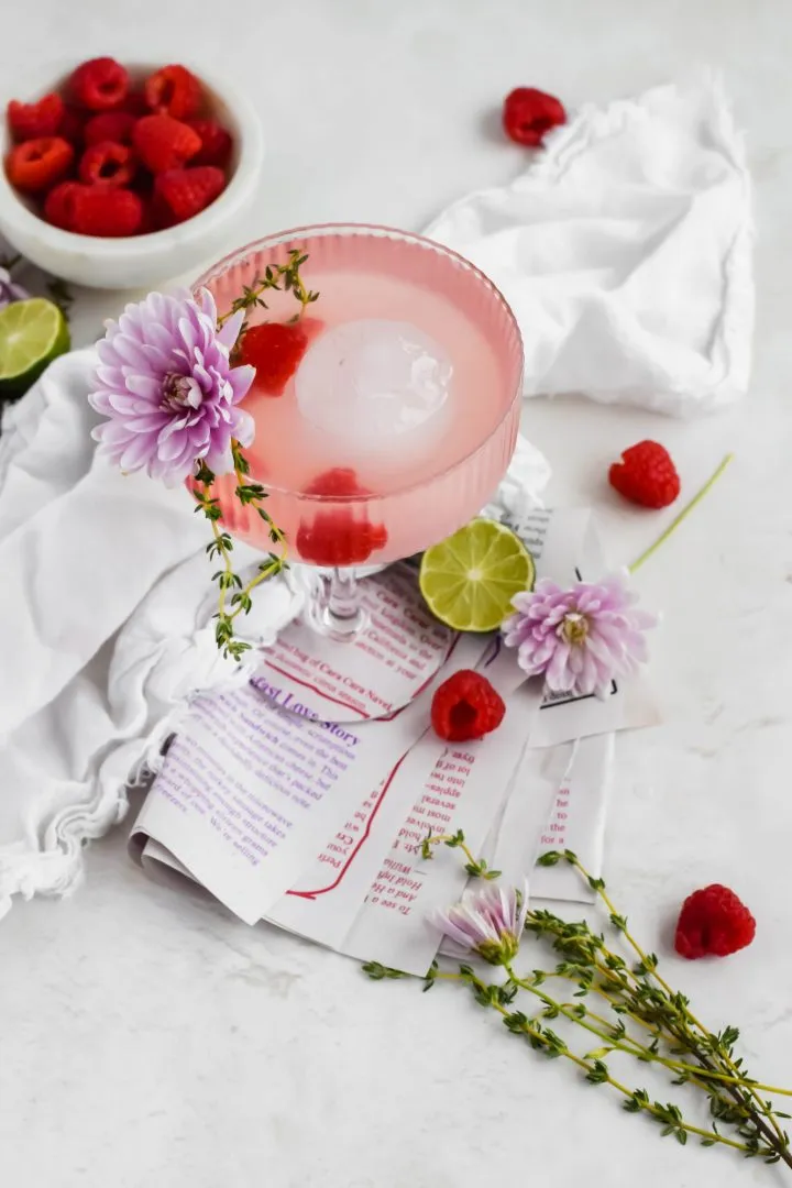 A pink cocktail in a glass, garnished with a fresh flower, sprigs of thyme, and fresh raspberries.