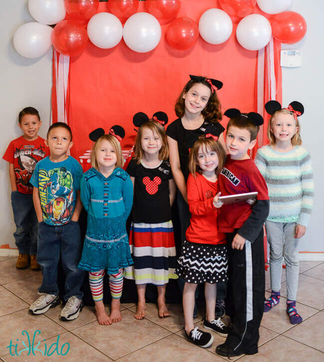 Eight kids wearing easy Mickey Mouse ears in front of a red and white party backdrop.