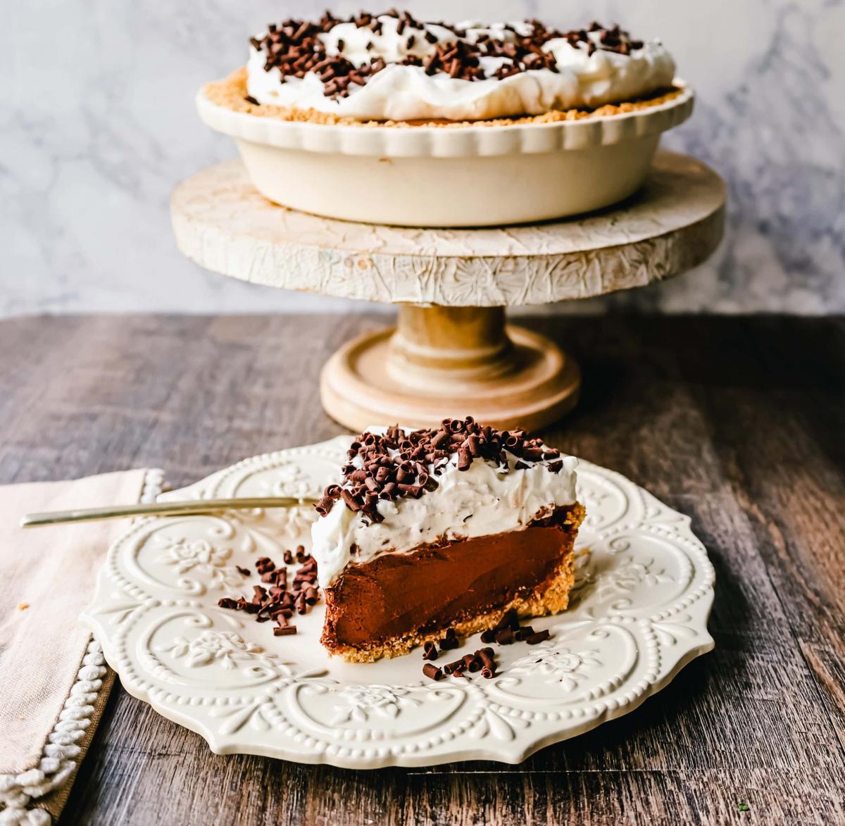 Slice of french silk pie topped with whipped cream and curls of chocolate on a white plate on a wooden surface.  The rest of the pie sits on a cake stand in a white pie dish behind the plate.