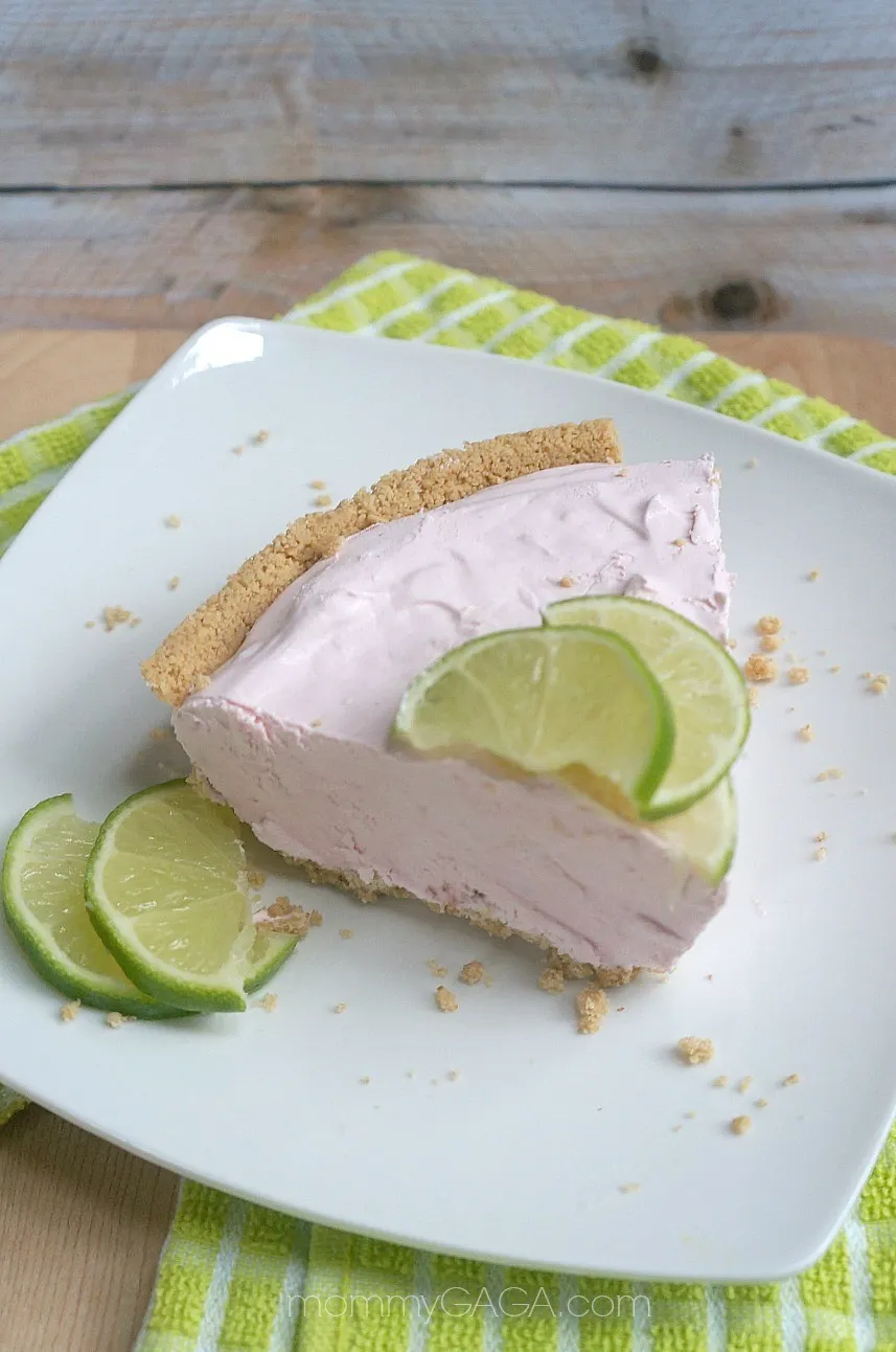 Slice of frozen cherry lime pie with a graham cracker crust on a white plate.  Slices of fresh lime garnish the pie.