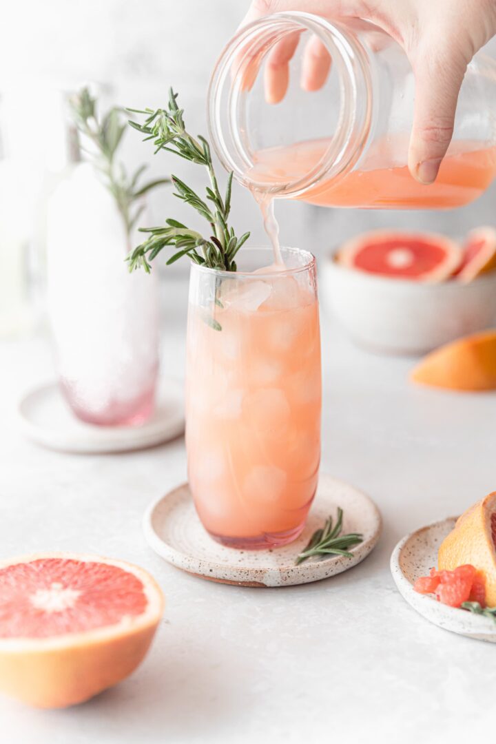 a blush pink grapefruit moscow mule cocktail being poured from a mason jar into a tall glass filled with ice and a sprig of fresh rosemary.
