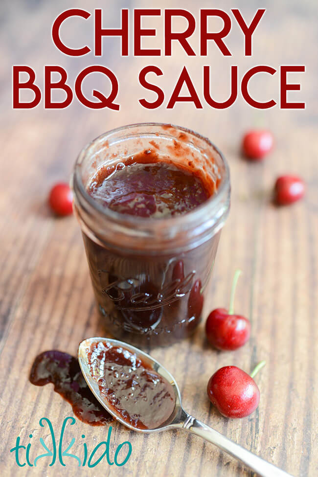 Open canning jar with cherry BBQ sauce, on a wooden table, with fresh cherries and a spoon full of cherry barbecue sauce on the table beside the jar.