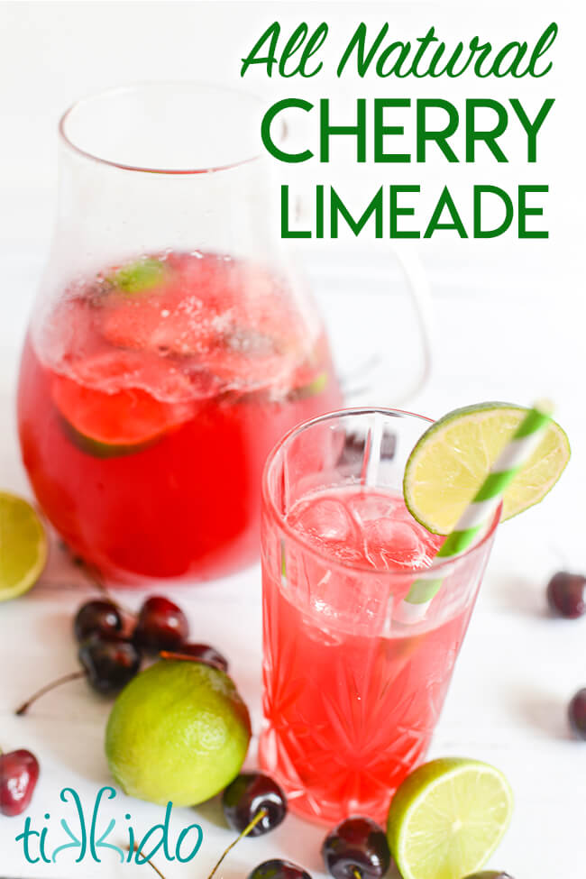 Glass of pink Cherry Limeade in a tall glass, with a green and white striped paper straw, garnished with a slice of lime.  The glass of limeade stands in front a pitcher of more cherry limeade, and fresh cherries and limes are scattered around the glass and pitcher.  Text overlay on the image reads "All Natural Cherry Limeade."