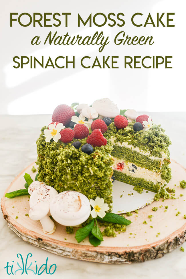 A green cake that looks like it is covered with moss, berries, edible flowers, and meringue mushrooms on a wooden tree slice, with text overlay reading "Forest Moss Cake, a Naturally Green Spinach Cake Recipe."