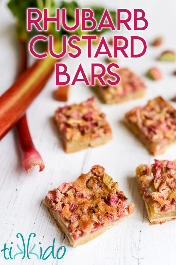 Five rhubarb custard bars on a white wooden surface, with fresh stalks and slices of rhubarb surrounding them, and text overlay reading "Rhubarb Custard Bars."