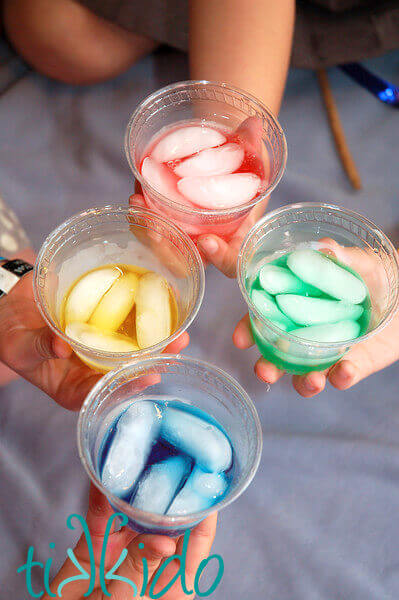 Four clear plastic cups filled with yellow, red, green, and blue liquid and ice, held by four different hands.