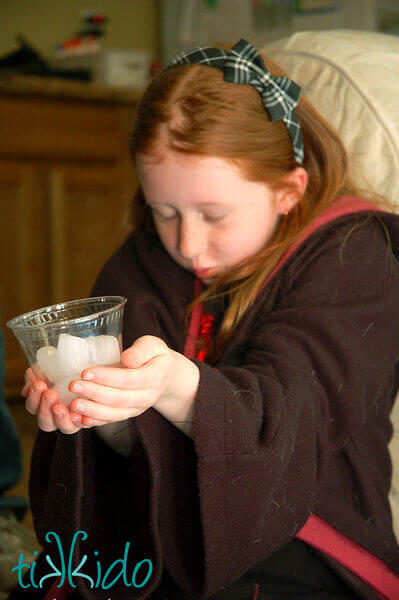 Red haired girl in Harry Potter robes, with closed eyes, holding a clear plastic glass with ice in her outstretched hands.