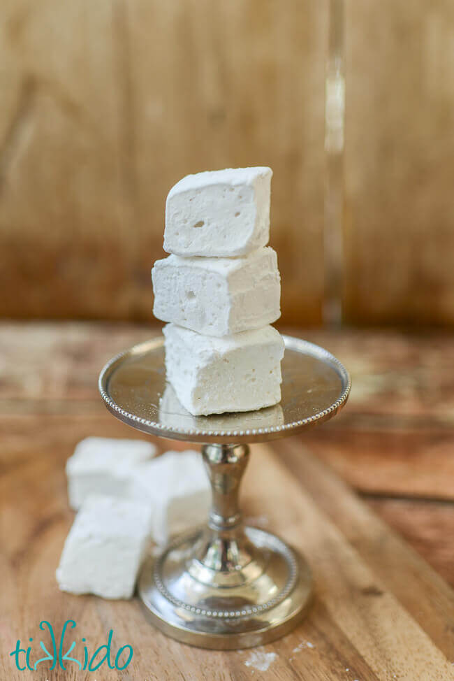 Three homemade marshmallows stacked on a tiny serving tray, and three more homemade marshmallows on the wooden surface below.