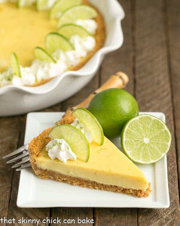 Slice of key lime pie garnished with lime slices and whipped cream, on a square white plate.  Two limes, one sliced in half, sit on the plate next to the pie and a fork sits behind the pie.