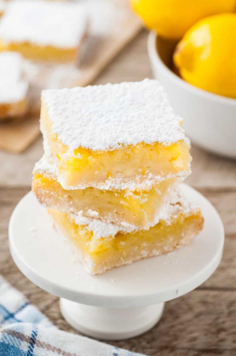 Lemon bars stacked on a small white cake stand, next to a bowl with fresh lemons.