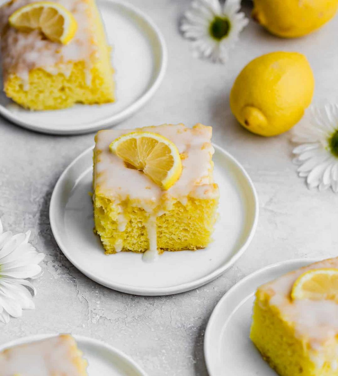 Slices of lemon jello poke cake on white dessert plates, surrounded by white daisies and whole lemons.