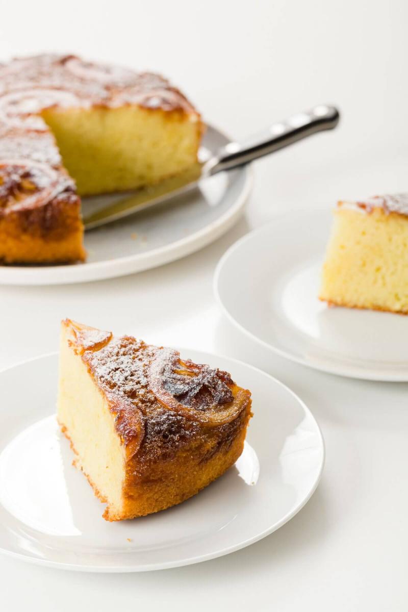 Slice of lemon olive oil cake on a white plate, in front of the rest of the cake on a cake plate.