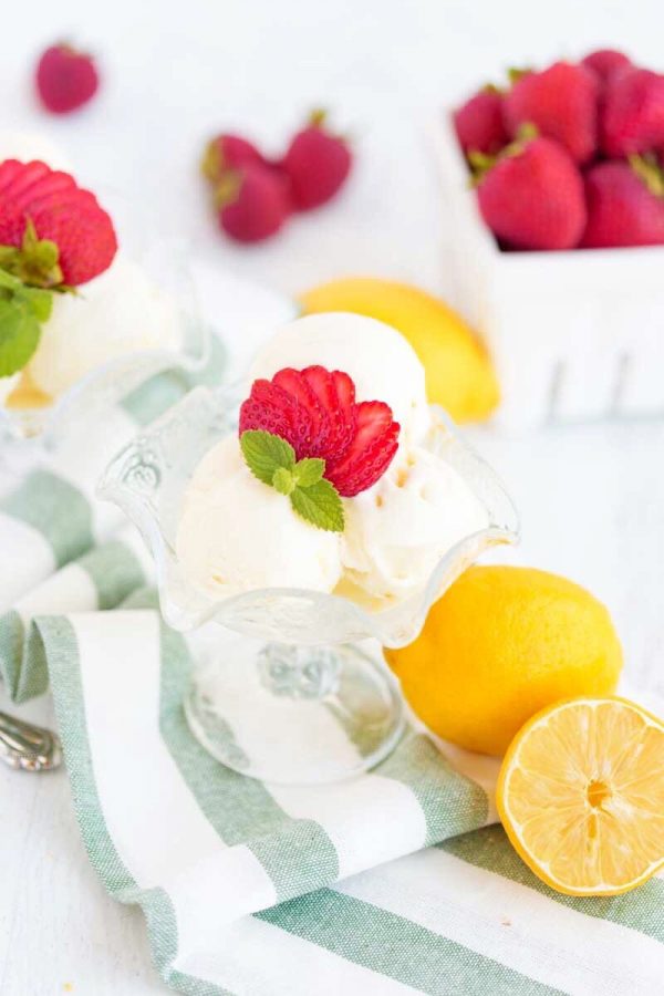 Scoops of lemon sherbet topped with sliced strawberry in a glass ice cream dish, next to whole lemons and strawberries.