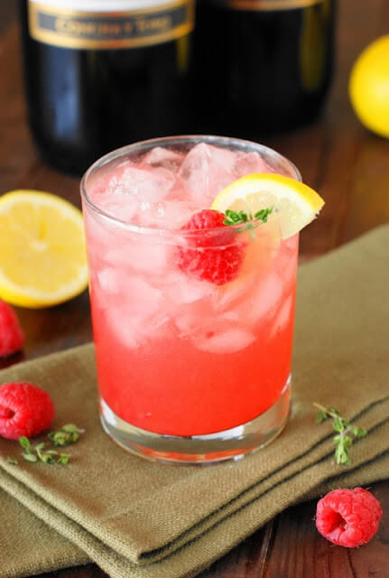 Pink Moscato Honey Bee cocktail in a lowball glass filled with ice, and garnished with a slice of lemon, a sprig of thyme, and a raspberry.