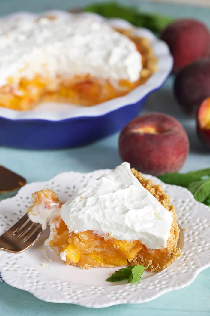 Slice of no bake peach pie on a white plate in front of a blue pie plate containing the rest of the pie.