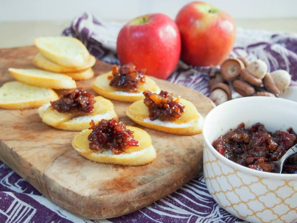 Apple bacon jam crostini on a wooden serving tray, surrounded by fresh apples and a dish of apple bacon jam.