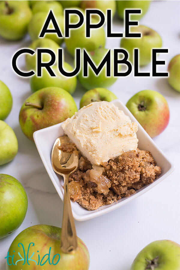 Apple crumble in a white bowl with a scoop of vanilla ice cream and a gold spoon, surrounded by green apples on a white marble background.