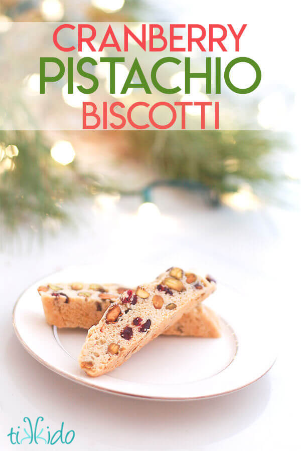 Two pieces of Cranberry Pistachio Biscotti on a white plate in front of Christmas lights and fresh greens, with text overlay reading "Cranberry Pistachio Biscotti."