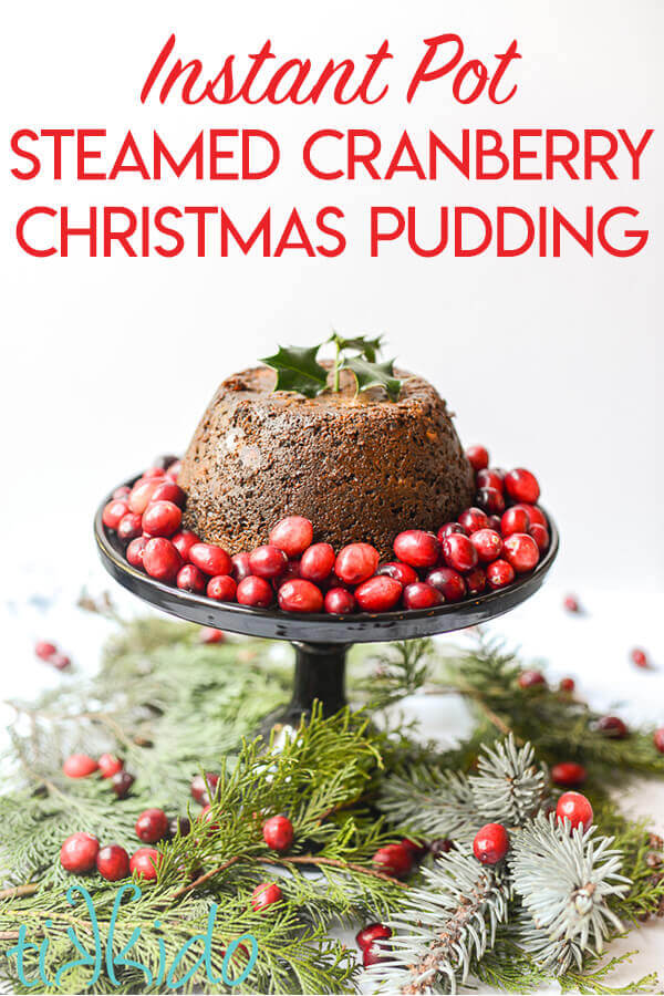 Cranberry Christmas pudding steamed in the Instant Pot on a cake stand, surrounded by cranberries and Christmas greenery.