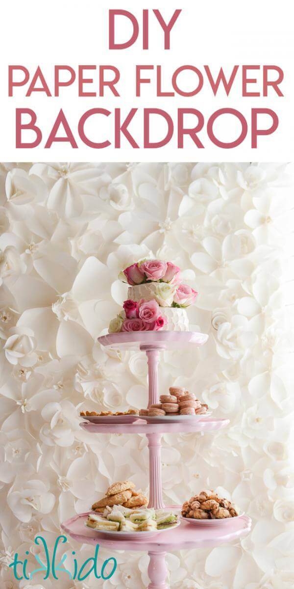 Monochromatic white paper flower backdrop behind a three tiered pink table with a cake on the top layer and tea party sandwiches and cookies on the lower layers.