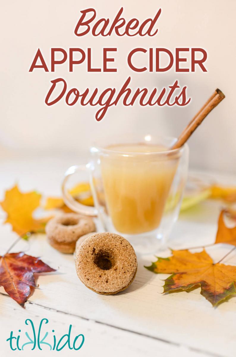 Miniature apple cider donuts next to a glass mug filled with fresh apple cider, with fall leaves scattered around the white wooden surface.  Text overlay reads "Baked Apple Cider Doughnuts."