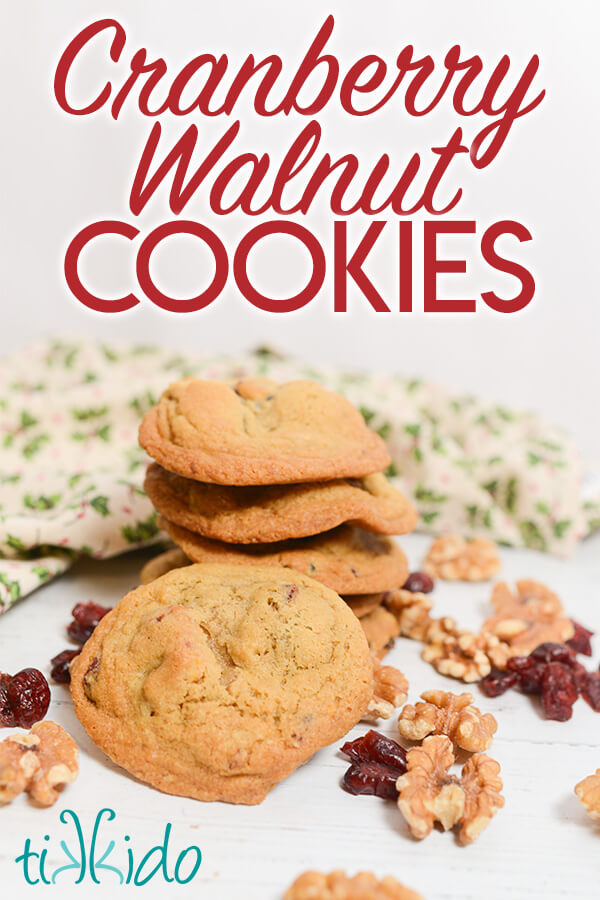 Stack of cookies surrounded by walnut halves and dried cranberries and a christmas holly patterned  tea towel on a white background.  Text overlay reads "Cranberry Walnut Cookies."