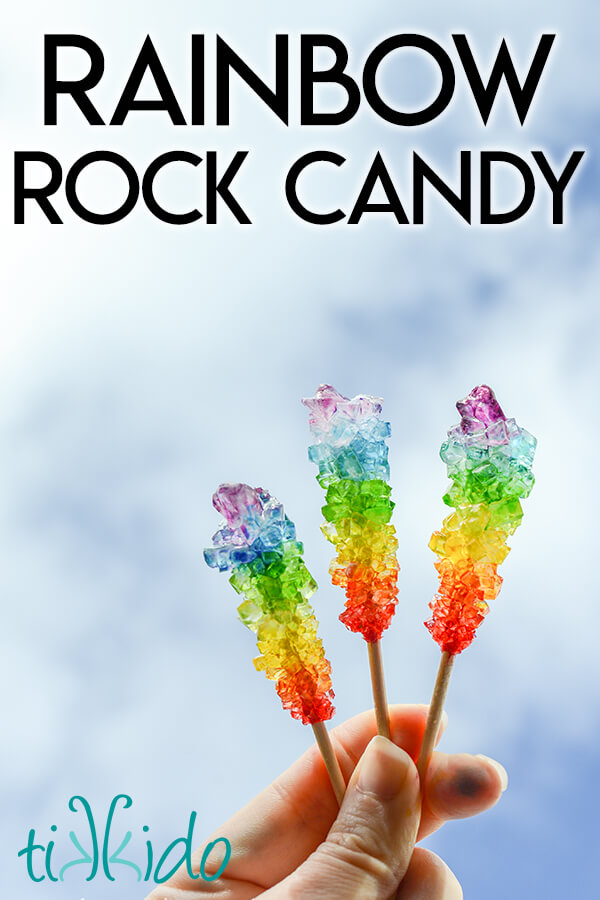 Three sticks of rainbow rock candy held up to a blue sky with white clouds.