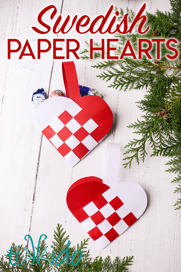 Two Swedish Paper Heart ornaments on a white wooden surface, surrounded by fresh evergreen branches.