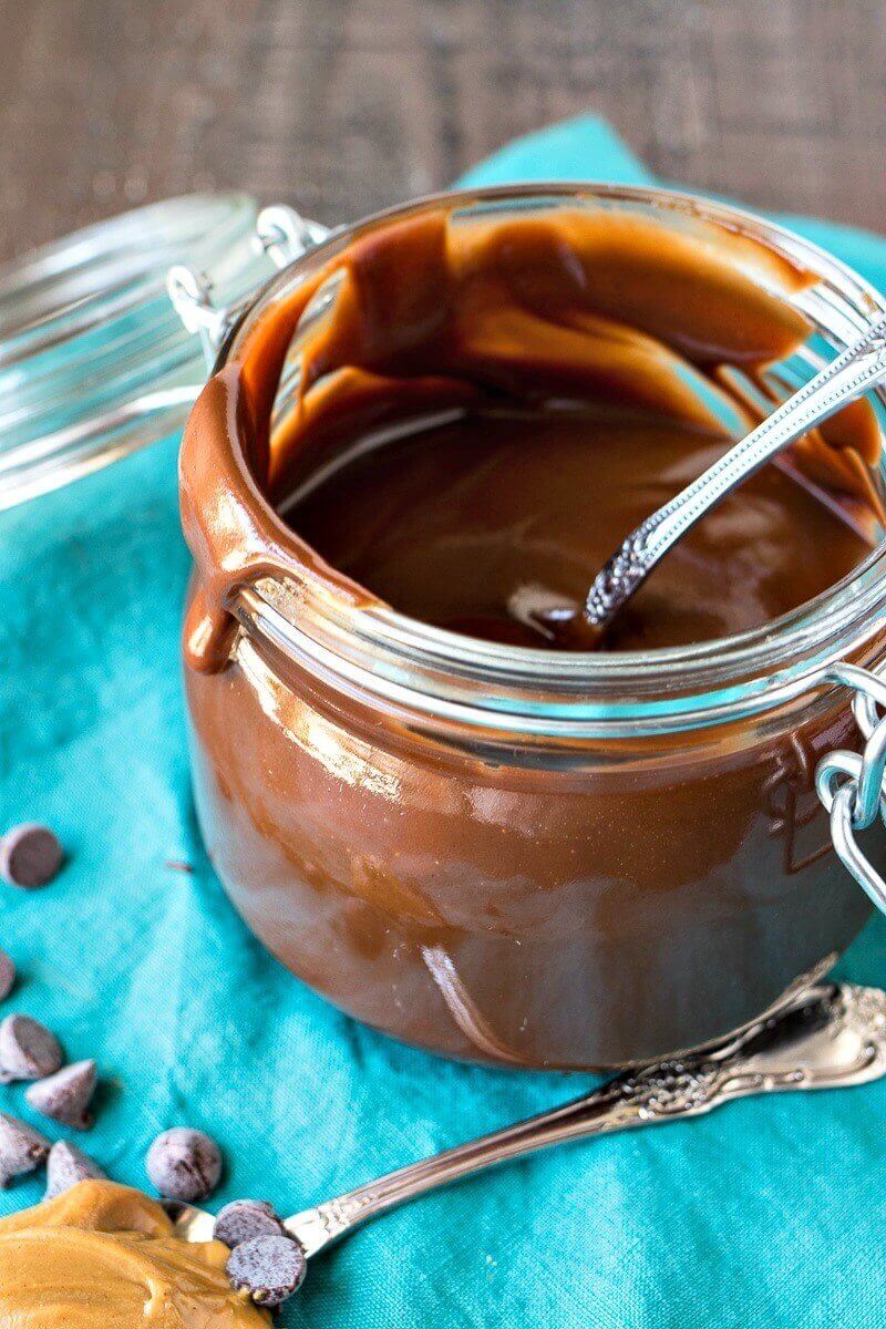 Glass pot of peanut butter hot fudge with a silver spoon, on a blue cloth.