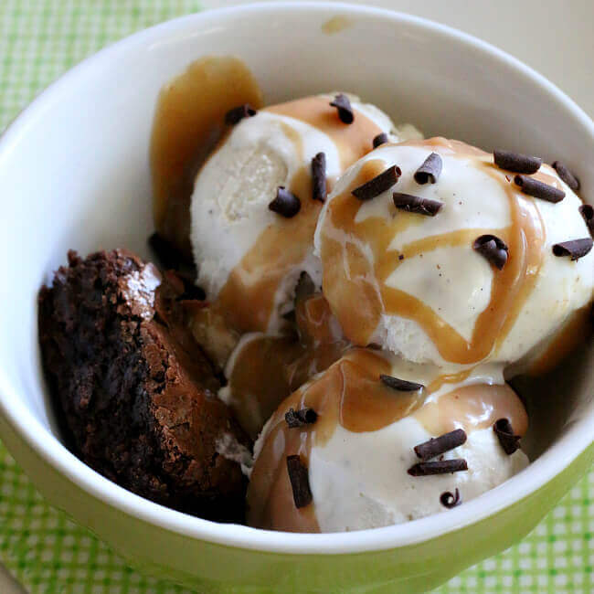 Peanut butter ice cream topping poured over vanilla ice cream and a brownie, in a white bowl, topped with curls of chocolate.