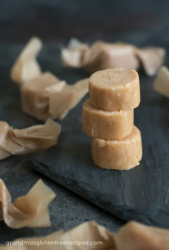 Old Fashioned peanut butter candy stacked on a slate surface, surrounded by homemade candies wrapped in waxed paper.