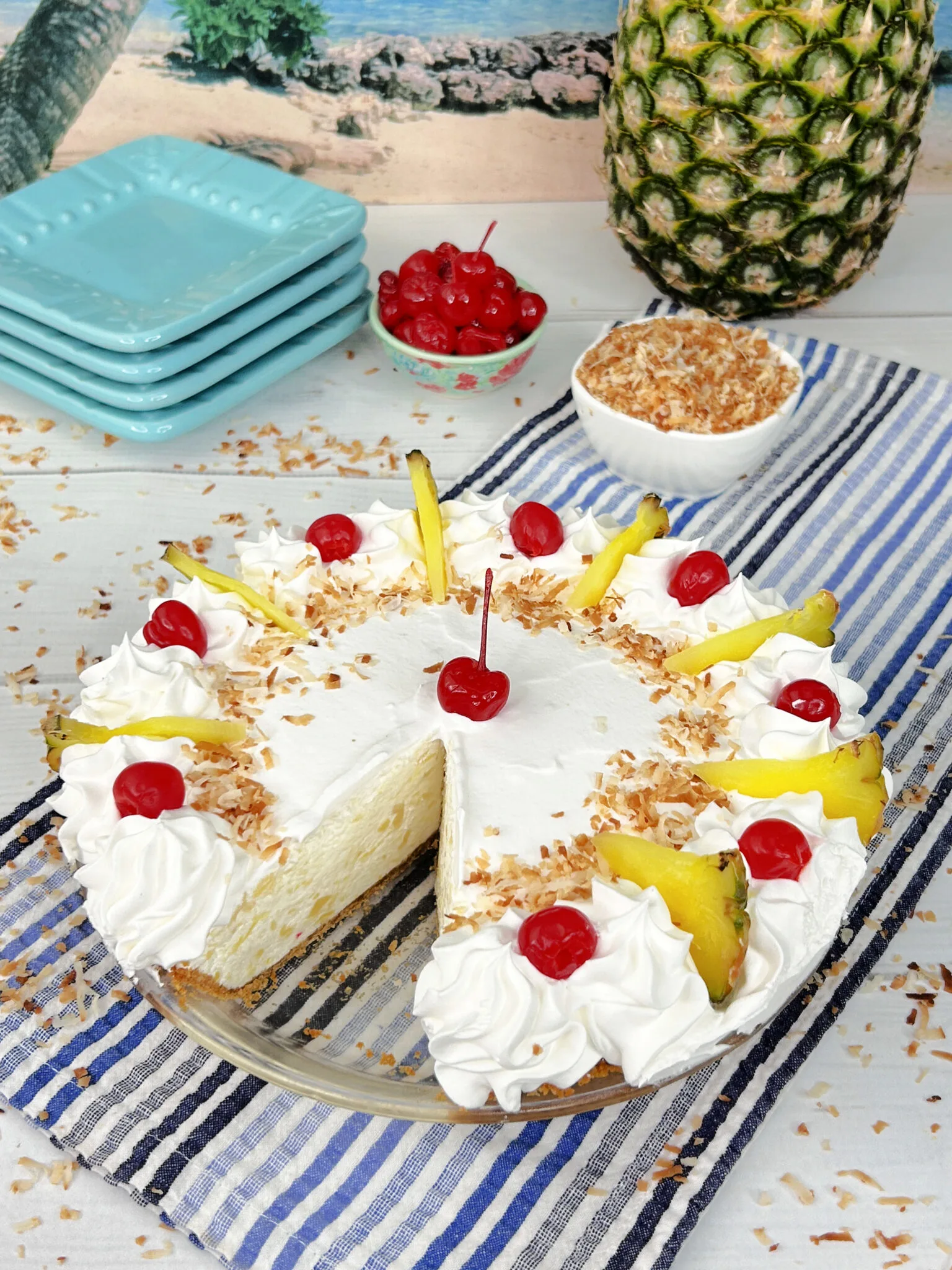 Pina colada pie with one slice missing in a glass pie plate.  The pie is garnished with whipped cream, toasted coconut, slices of pineapple, and maraschino cherries.