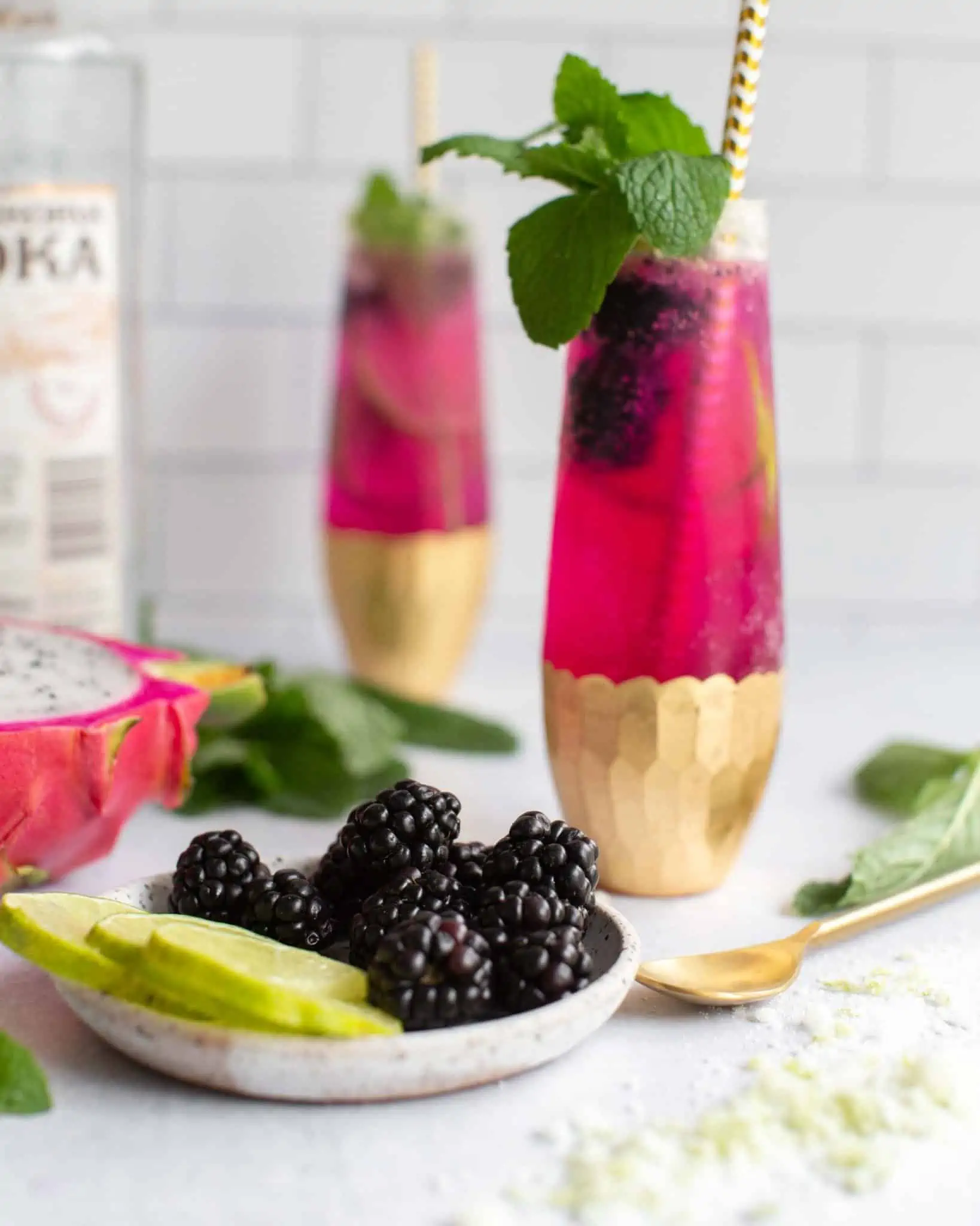 A dark pink dragonfruit cocktail in a tall glass painted gold at the bottom, garnished with fresh blackberries, sprigs of mint, and a gold and white paper straw.