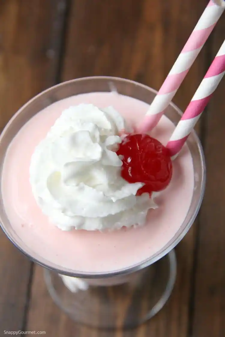 Pink squirrel cocktail topped with whipped cream and a maraschino cherry, with two pink and white paper straws sticking out of the glass.