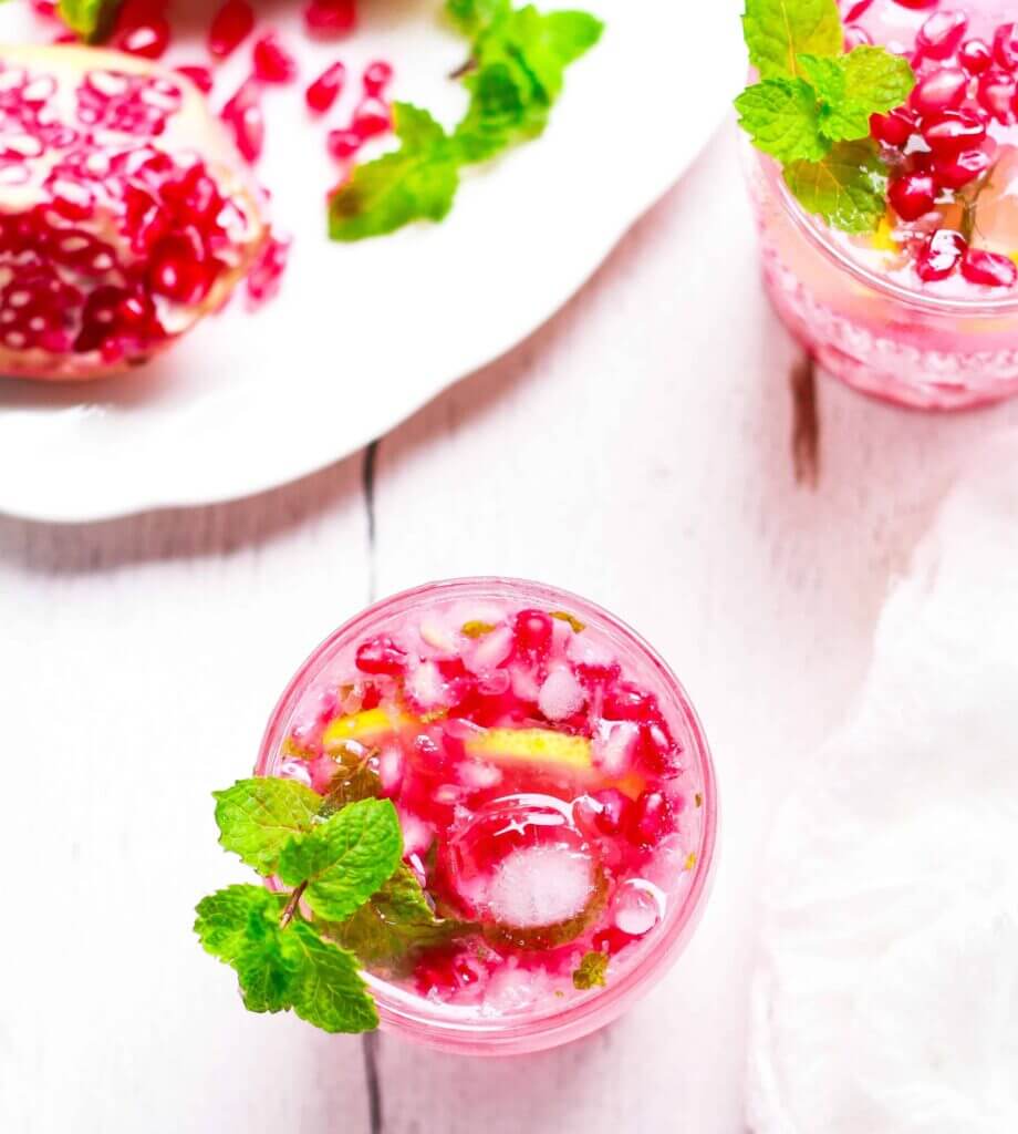 two glasses of bright pink pomegranate moito garnished with sprigs of fresh mint on a white wooden surface.  Half a pomegranate and pomegranate seeds sit on a white plate next to the glasses. 