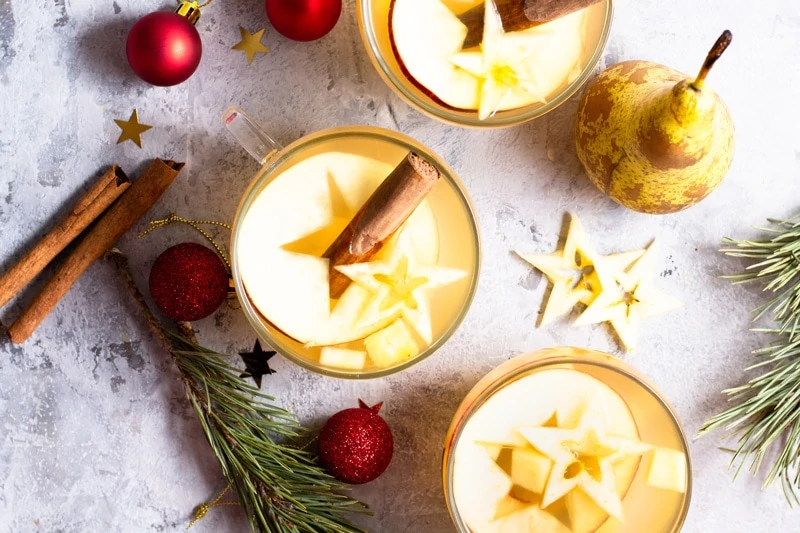 Three glasses of Ponche de Frutas, a latin American spiced Christmas punch, on a grey surface, surrounded by fresh evergreen branches, red Christmas ornaments, and cinnamon sticks.