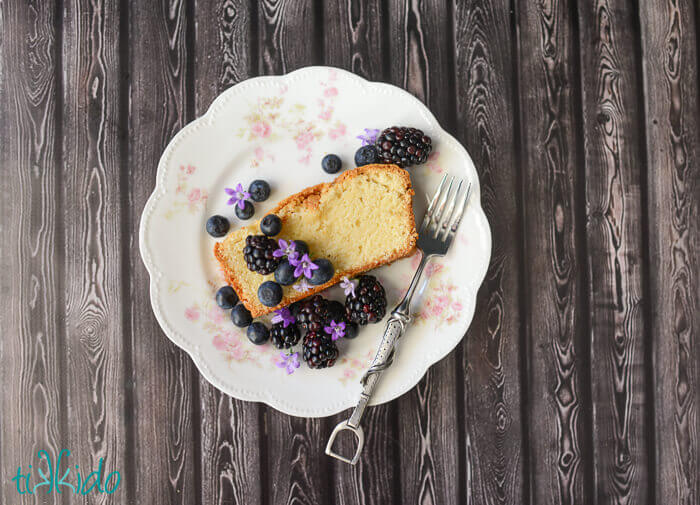 Slice of pound cake topped with blackberries, blueberries, and edible purple flowers, on a floral plate with a fork on a dark background.