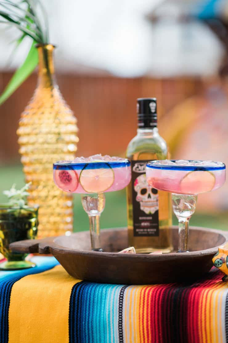 Two margarita glassis filled with pain pink prickly pear margaritas, in a wooden tray sitting on a colorful striped Mexican blanket.  A bottle of tequila sits in the background.
