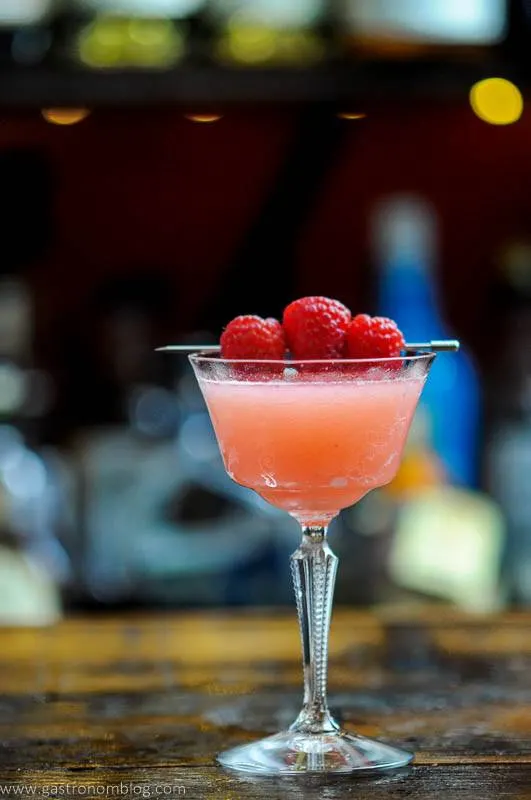 Raspberry gimlet, a pink version of the classic gin cocktail, garnished with three fresh raspberries, sitting on a wooden table.