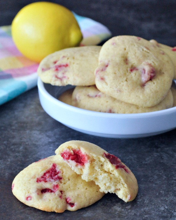 Raspberry lemon vegan cookies in a white dish, with two cookies on the table in front of the dish.  One of the cookies is broken in half so you can see the raspberries inside and soft texture.
